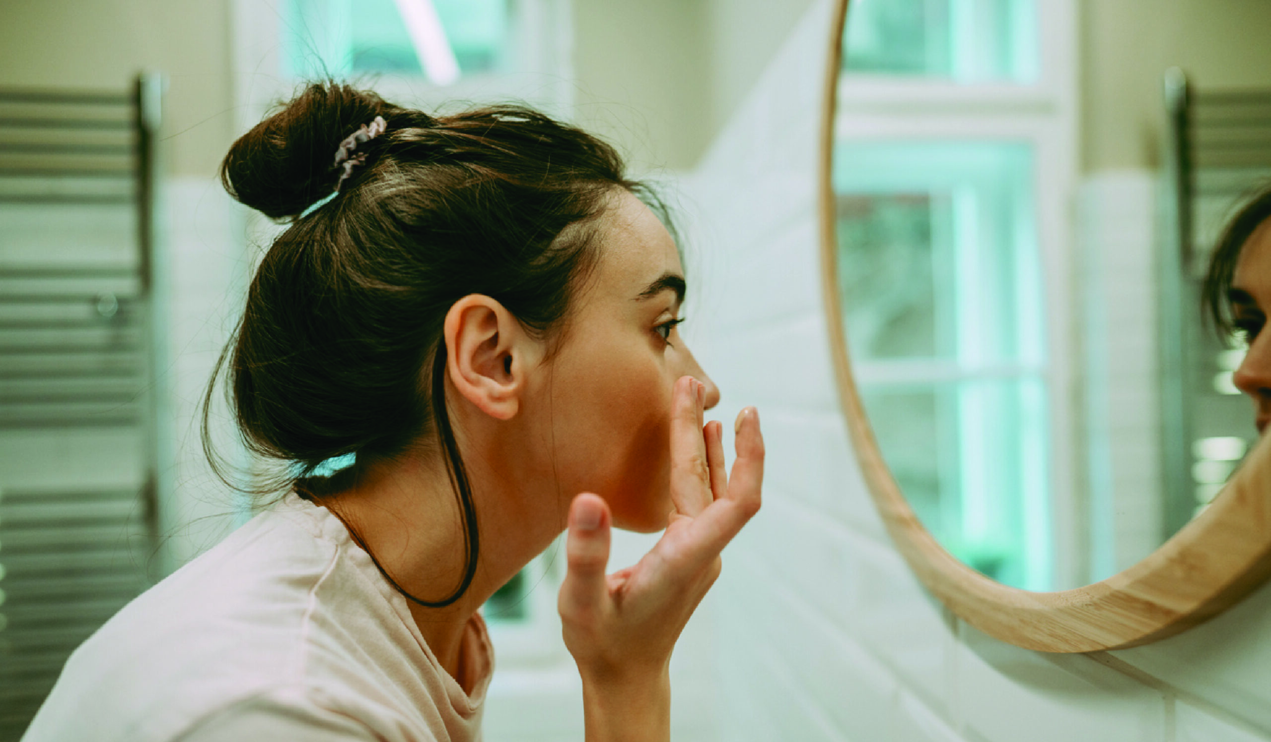 Woman looking in mirror while gently massaging her face with a skincare product. She appears relaxed and focused, engaging in a self-care routine.